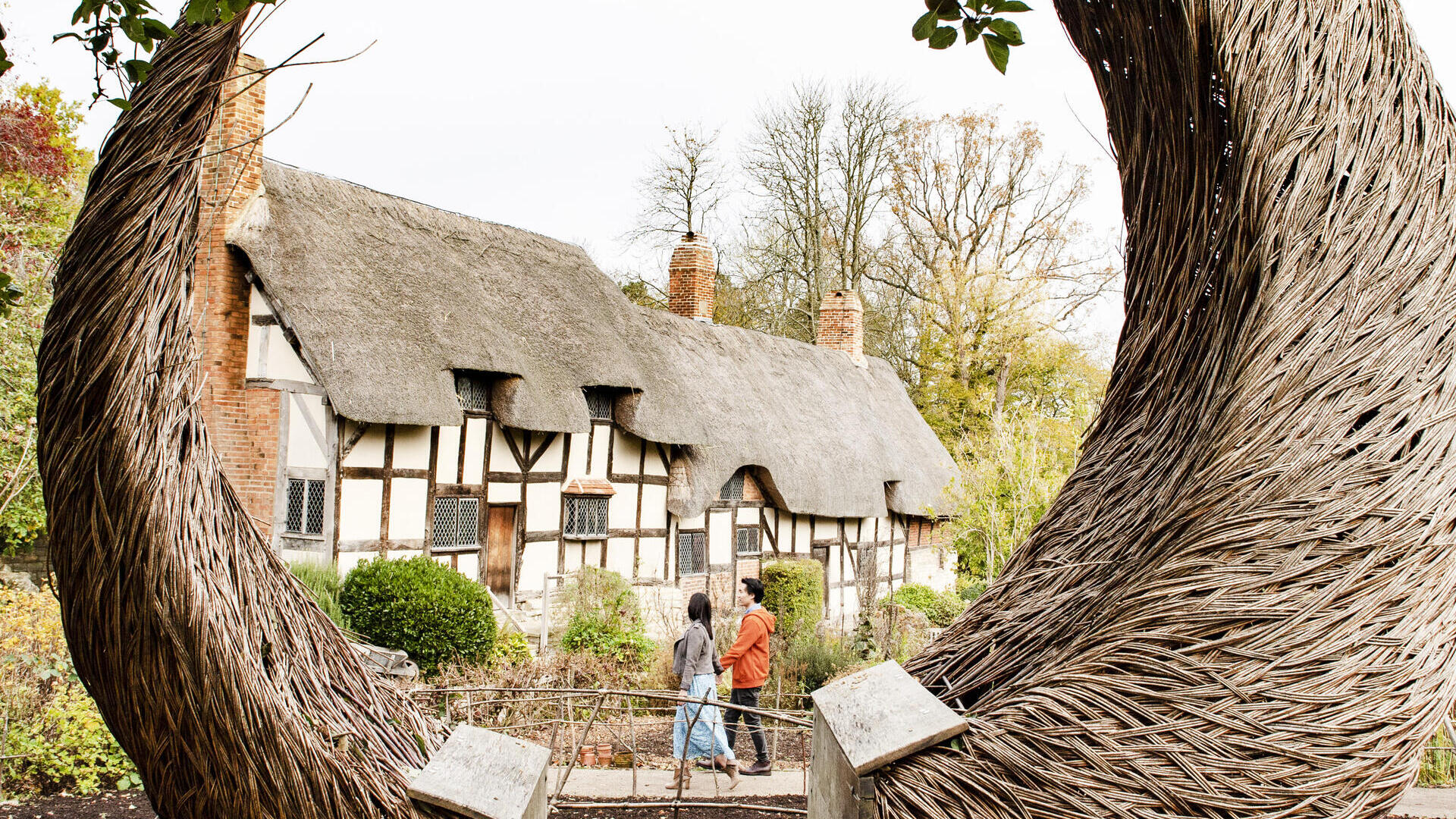 Couple walking near a cottage seen through a large willow crescent shaped sculpture in a garden