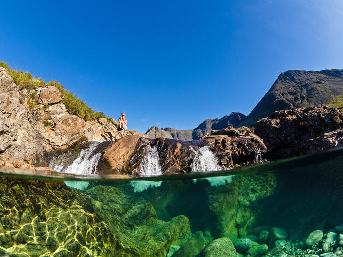 Clear stream with visible underwater rocks, small waterfall, rocky landscape and mountains under a blue sky. Person sits on rocks above the water.