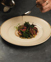 Plated gourmet dish being garnished with sauce, featuring sliced meat, greens, and a creamy puree on a patterned plate.