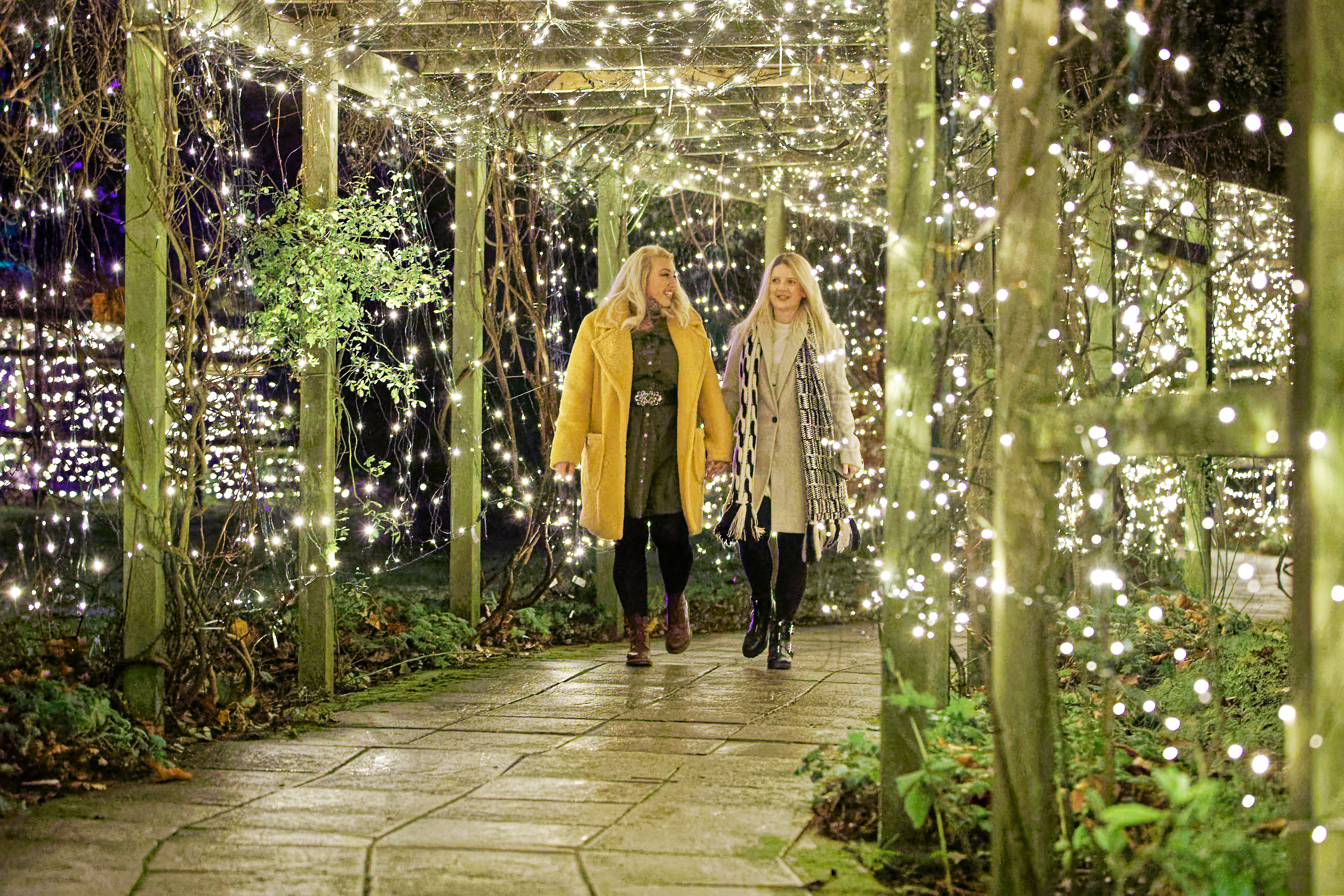 Two women walking together through a lit walkway covered in plants.