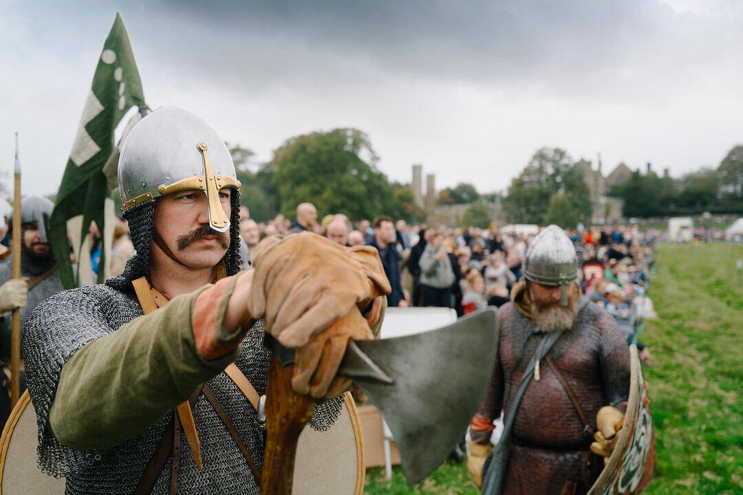 Historical reenactors dressed in medieval armor with weapons, performing before a crowd in an outdoor setting with historic buildings.