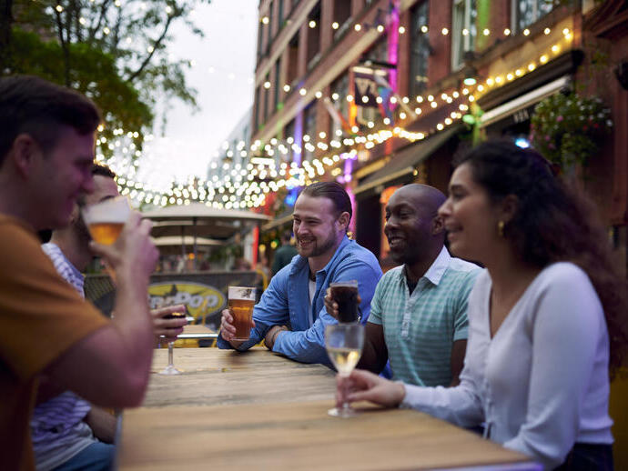 Freunde, die an einem Tisch im Außenbereich einer Bar mit Lichterketten im Hintergrund etwas trinken