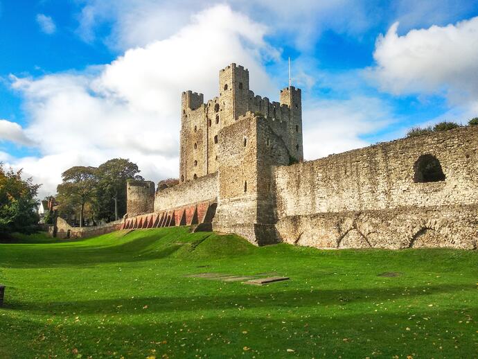 Large stone castle with fortified walls, towers, and green lawn under a bright blue sky with scattered clouds.