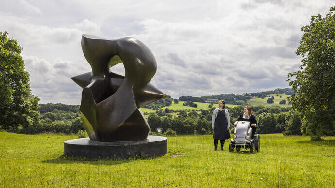 Two women look at a large sculpture set in green gardens with landscape beyond