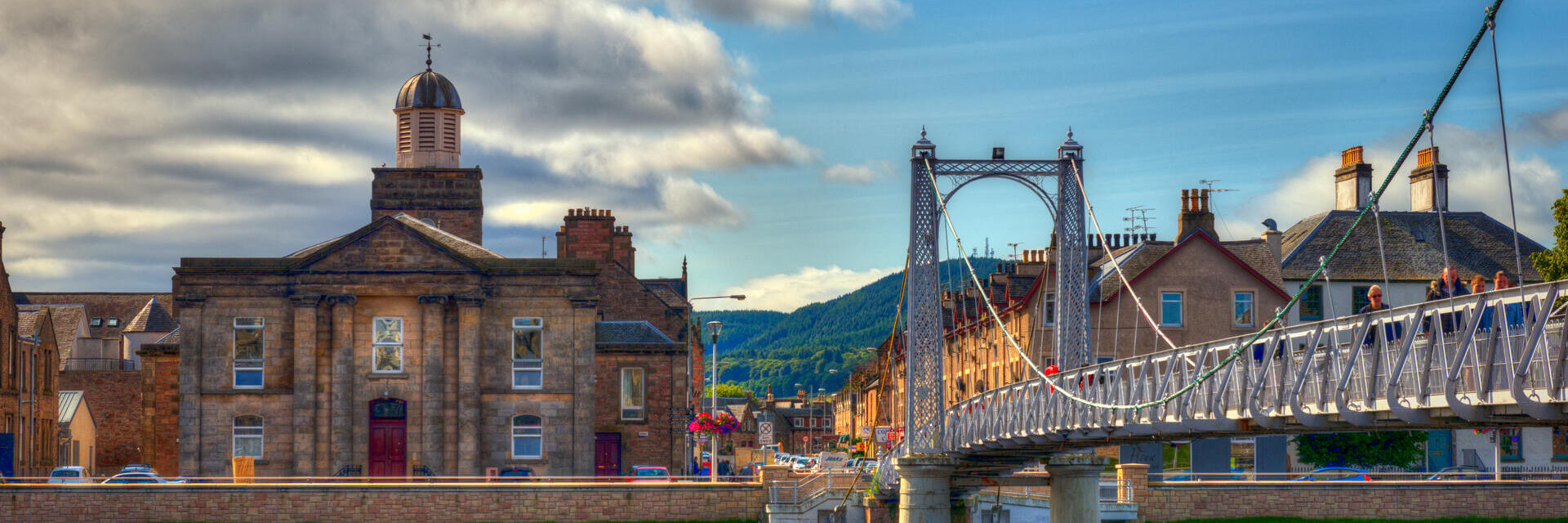 A suspension bridge with people walking across spanning a wide river with houses on the other side