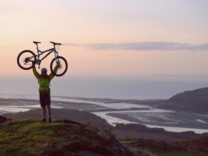 Man holding his mountain bike above his head in celebration at the top of a hill with the coast below him