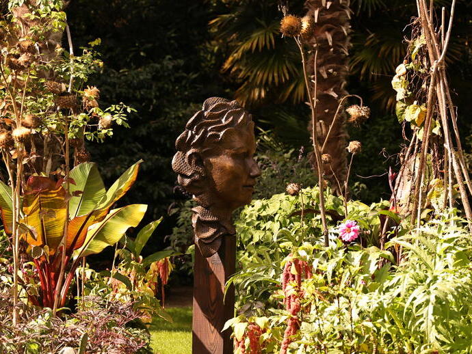 A bronze head sculpture in a lush garden surrounded by green foliage, tall plants, and flowers in sunlight.