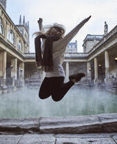 Frau springt in die Luft in einem römischen Freiluftbad, umgeben von historischen Gebäuden