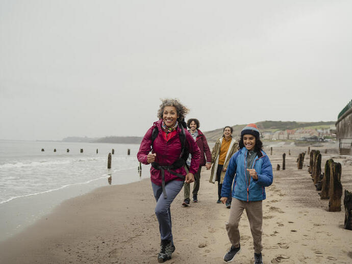 Group of people hiking and smiling along a sandy beach with wooden groynes, some houses, cliffs, and sea in the background on a cloudy day.