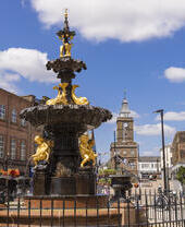 An ornate black and gold fountain with statues of people and fish in the centre of a shopping street.