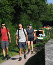 Three people walking on a path along a canal with boats moored.