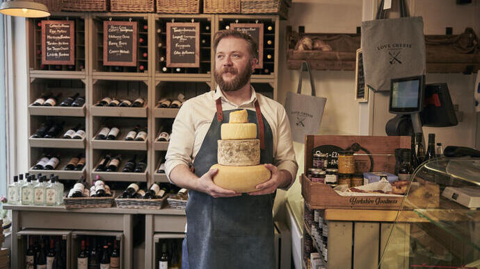 Man wearing blue apron holding a stack of cheeses in a cheese and wine shop