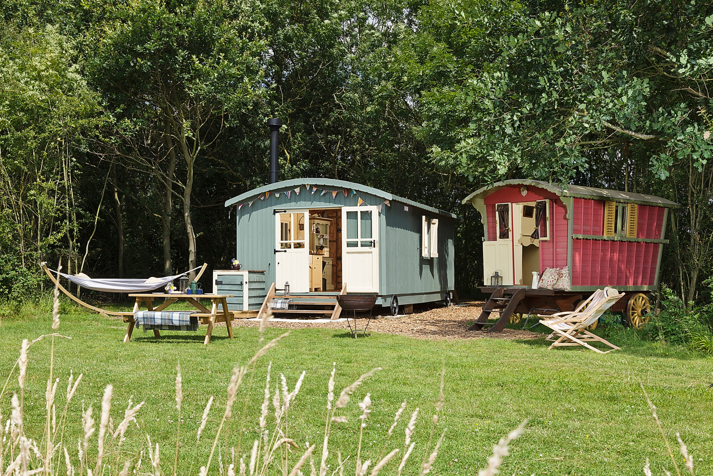 Colourful shepherd's hut and gypsy caravan style accommodation in a tree-edge field with a hammock and picnic table in front