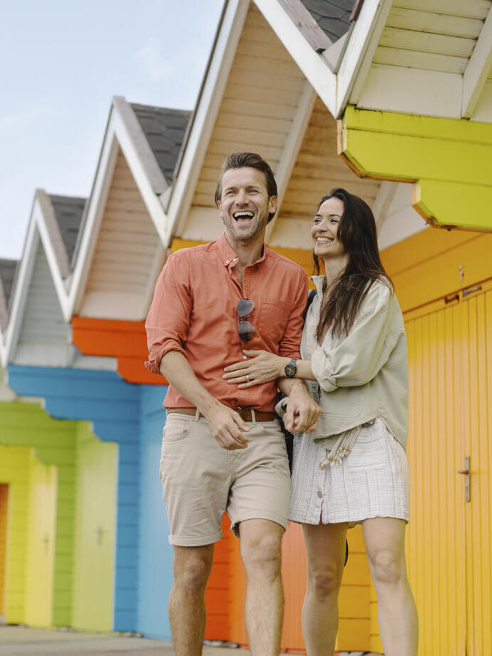 A man and woman laughing outside colourful beach huts
