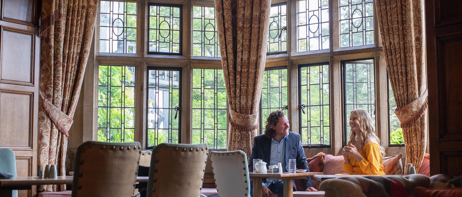 A man and a woman have coffee in a hotel lounge with large windows.