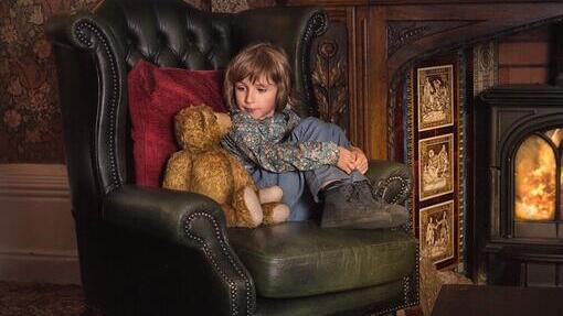 Young child sat on a green armchair in front of a fireplace with a teddy bear next to them at Criag y Nos Castle in the Brecon Beacons