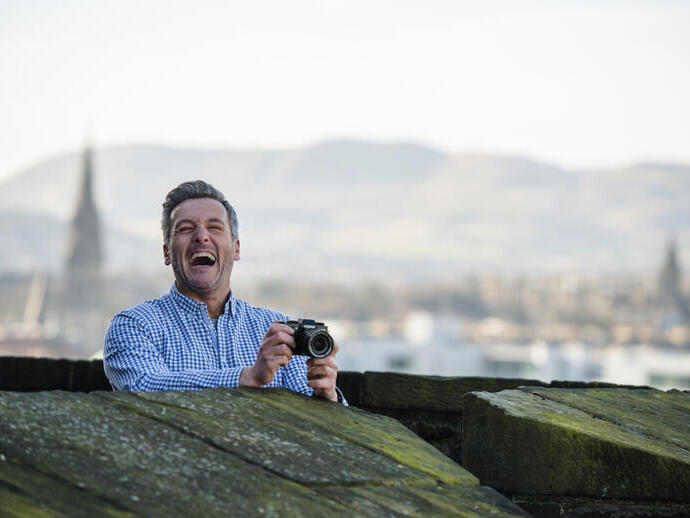 Smiling person holding a camera stands behind stone wall, with blurred cityscape and hills in the background.