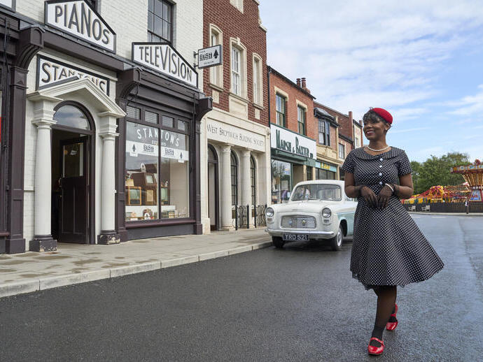 Woman in 1950s-style dress walks past vintage shops, classic car, and fairground ride on a recreated historic street scene.