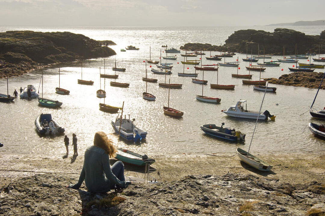 A woman looking over boats in a village harbour at sunset.