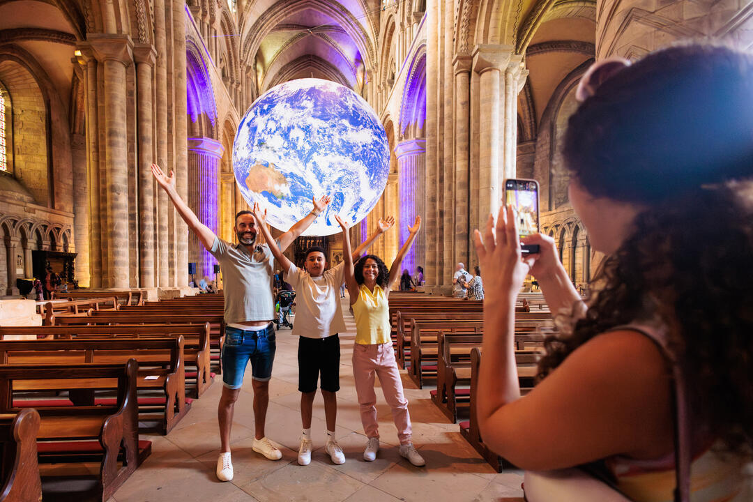 A family take a photo in front of an installation of a large hanging globe inside a cathedral
