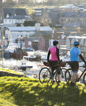 Two cyclists with gear overlook a harbor with boats and historic stone buildings under sunlight, suggesting outdoor adventure and scenic town views.