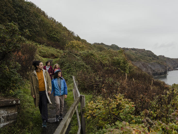 Group walking along a coastal path with lush greenery, cliffs, and sea in the background on a cloudy day.