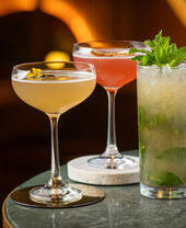 Three cocktails on a marble table: two in coupe glasses with garnishes and one tall mojito with mint leaves, set against a warm background.
