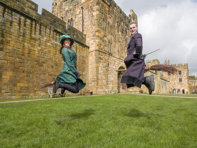 Two wizarding professors demonstrating flying on a broom outside a large castle.