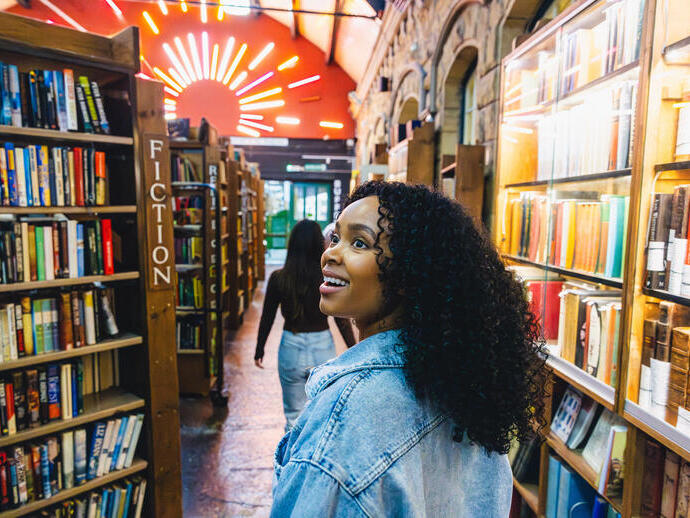 Two people walk through a vibrant, well-lit bookstore with shelves of books, stone walls, and neon lights overhead.