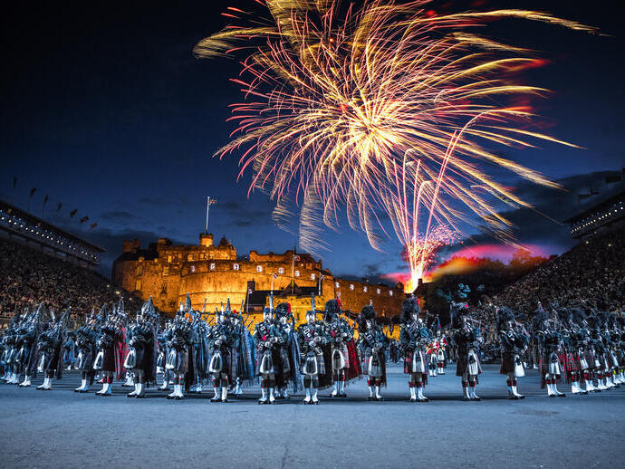La arena del Edinburgh Military Tattoo durante una actuación del evento militar, el patio de armas y las gradas repletas de espectadores. Un espectáculo de luces proyectado sobre las murallas del castillo. Banda de música con un director al frente y un grupo de gaiteros tocando la gaita. Fuegos artificiales explotando en el cielo nocturno.