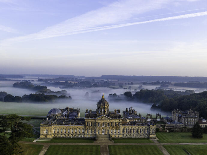 Grande maison de campagne vue d'en haut avec des jardins à la française et une campagne boisée derrière, recouverte de nuages bas