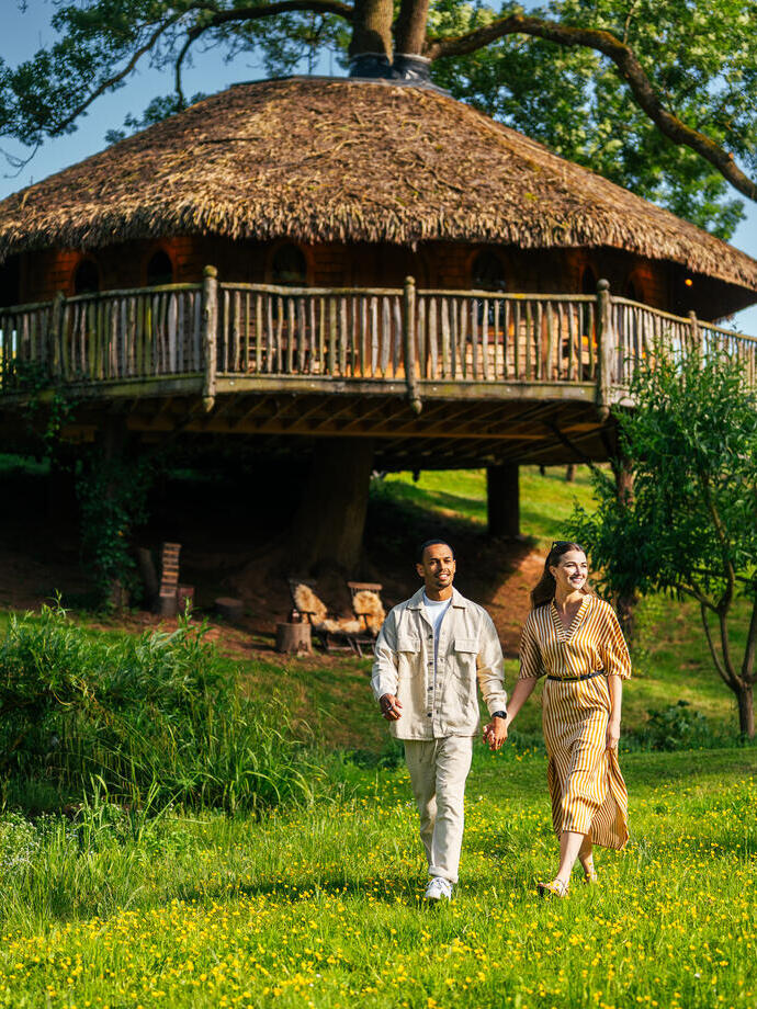 A man and a woman walk with a glass of wine in hand in the ground of Treetopia with the treehouse accommodation in the background