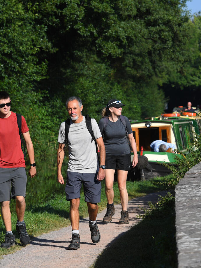 Three people walking on a path along a canal with boats moored.