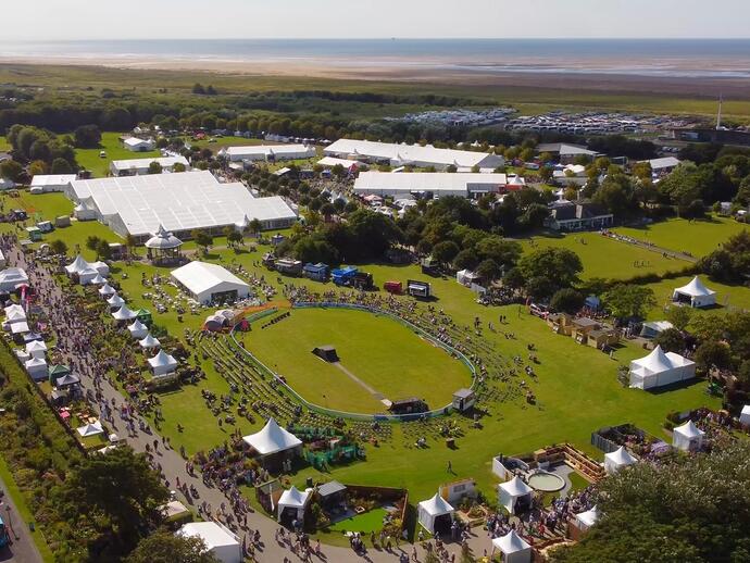 Aerial view of a large outdoor festival with crowds, tents, exhibition stands, event rings, lawns, and coastline in the background.