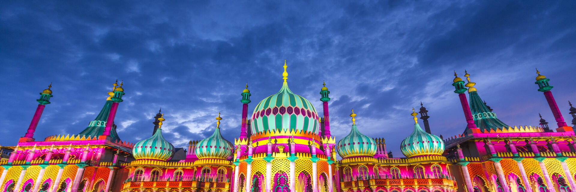 Multitud sentada en el suelo frente a un gran edificio ornamentado iluminado con colores vivos