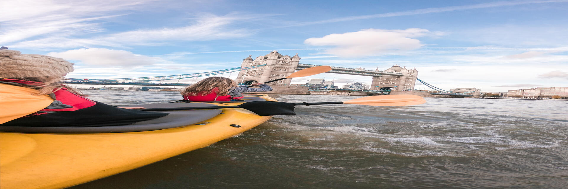 Two women paddling in a kayak on a river towards a bridge in a city