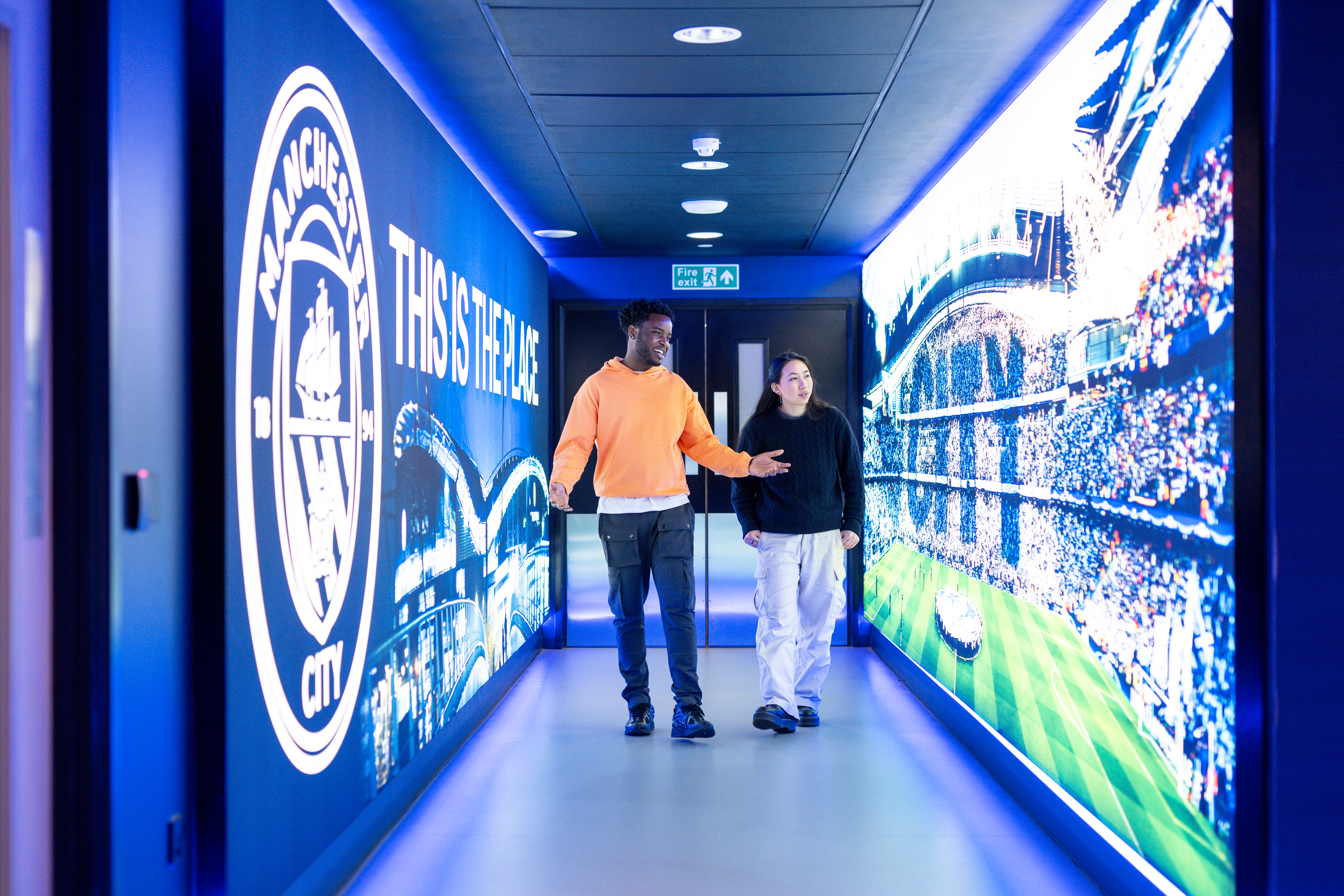 Two people walk through a modern hallway with bright displays, including a football stadium and Manchester-themed graphics.