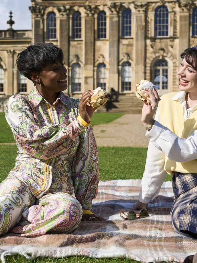 Two people enjoy a picnic on a blanket in front of a grand historic building with large windows and columns, on a sunny day.