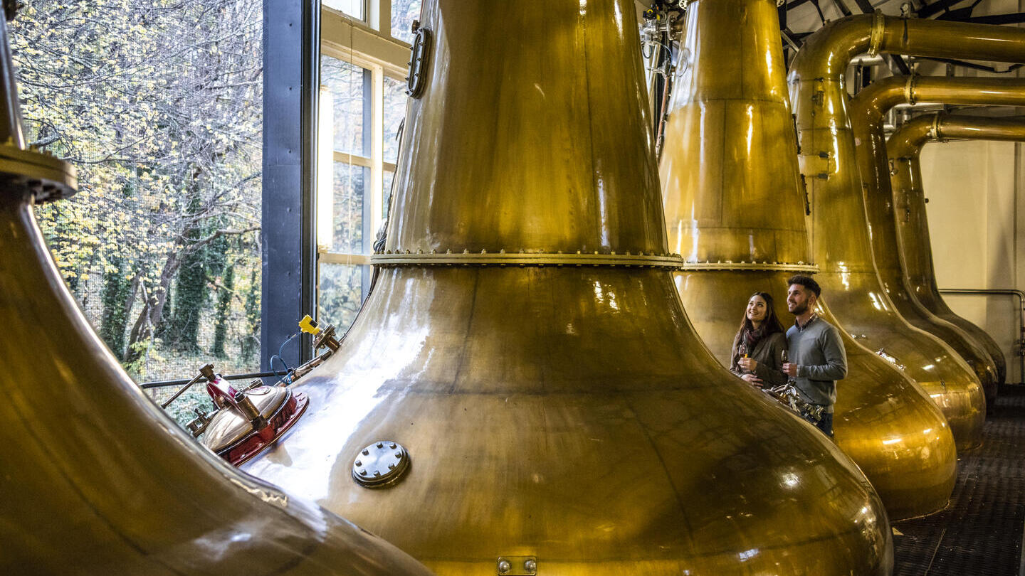 Pareja de pie junto a grandes alambiques de cobre en una destilería, bebiendo