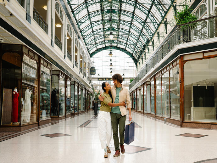 A couple walks inside a historic shopping arcade with a large glass domed roof, surrounded by elegant shopfronts and mannequins on display.