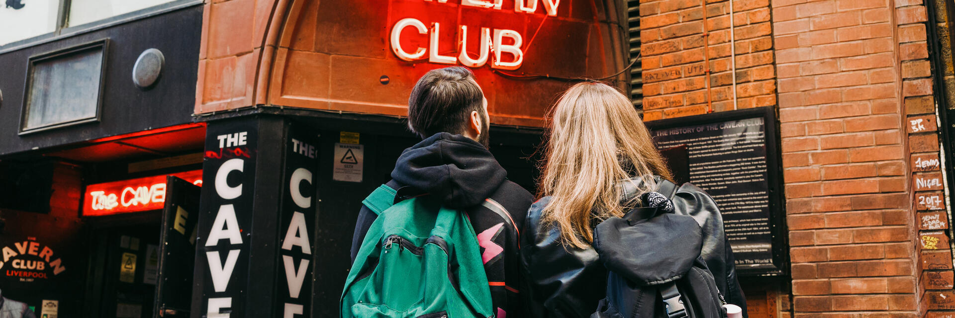 Due persone con zaini fuori dall'ingresso di un club con un'insegna al neon rossa illuminata.