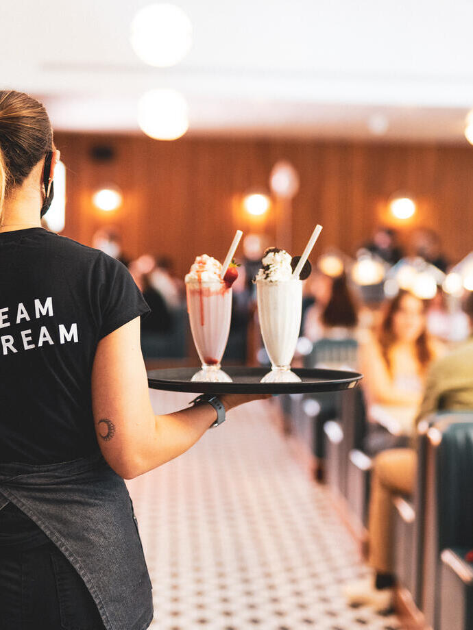 Staff member carrying two milkshakes on a tray in a diner.