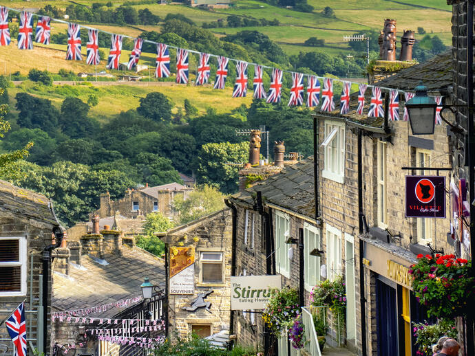 Gente caminando por una empinada calle empedrada de un pueblo con banderines de la Union Jack colgando de las casas