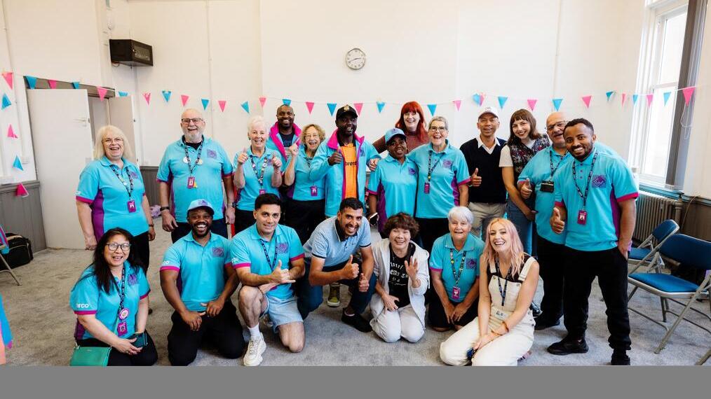 A diverse group of people in matching blue shirts gather in a decorated room, posing and smiling for a group photo, suggesting a team or community event.