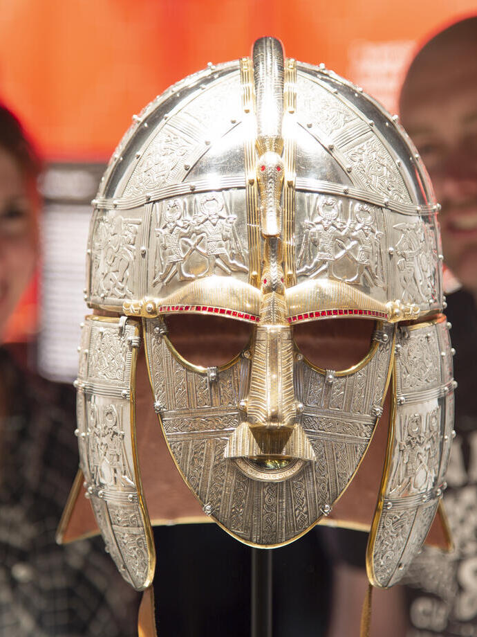 Two people admire an ornate Anglo-Saxon helmet in a museum display with a bright orange backdrop.