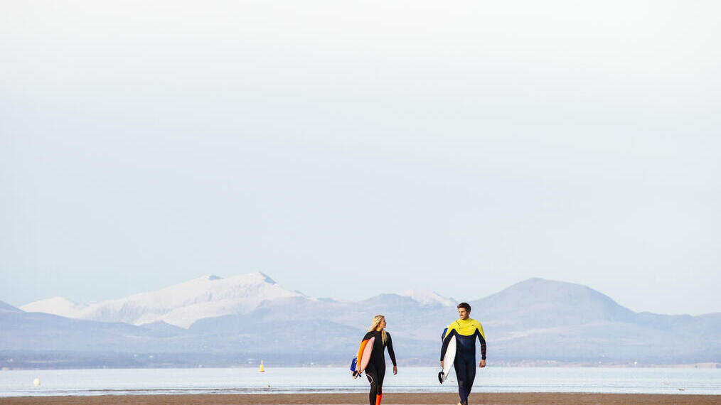 Man and woman, in wetsuits, carrying surfboards on beach
