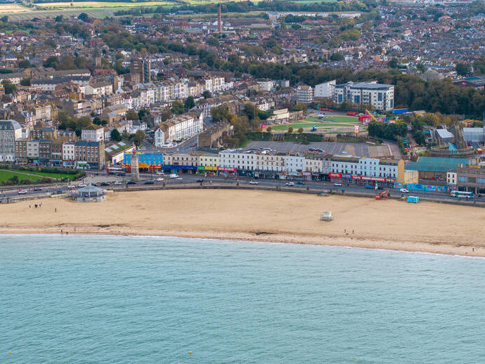 Aerial view of a sandy beach with a promenade, amusement rides, and a town with buildings and greenery in the background.