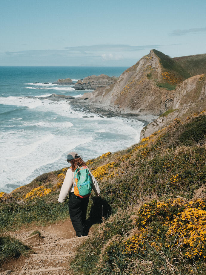 A woman walks along a coastal path with sea and headland in the distance