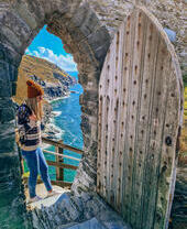 A woman stands at the top steps of a doorway with coastline view beyond