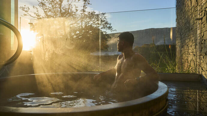 A man relaxes in a steaming outdoor hot tub as the sun rises, surrounded by nature and modern stone architecture.